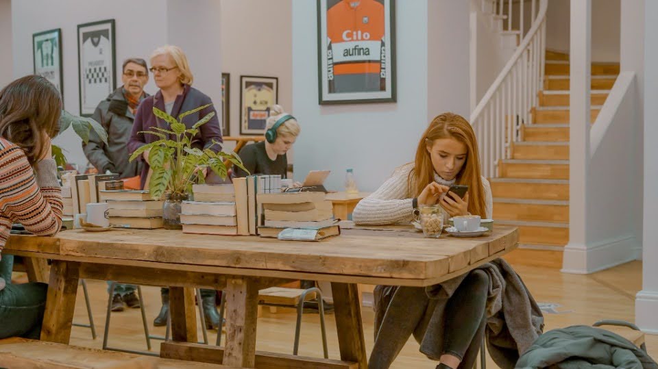 A woman checking her phone at a table stacked with books.