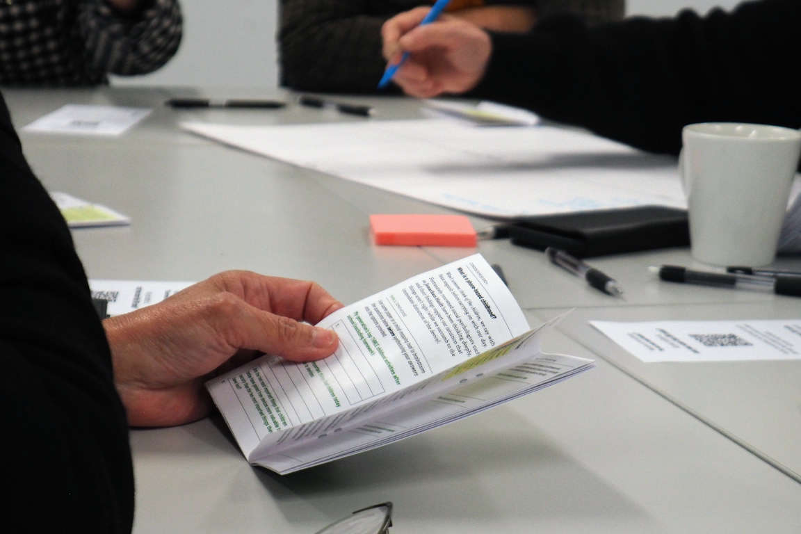 A hand holding a folded printed booklet on a table, with a QR-coded card nearby.